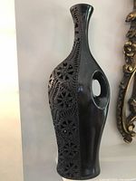 Full view of tall black barro negro pottery vase showing carved floral cut-out details on one side and open handle.