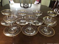Seven vintage clear glass champagne/sherbet glasses each paired with a matching plate, all with gold rim trim, displayed on wooden table.