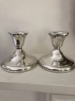 Photo showing two sterling silver weighted candle holders side by side, viewed from the front with reflective and worn surfaces visible.