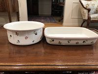 Two ceramic baking dishes on a wooden surface, round and rectangular, with small floral decorations and clean surfaces.