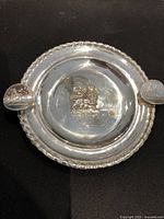 Full view of round silver ashtray dish with rope edge and two arms holding a coin each, central lion detail visible.
