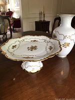 White pedestal cake plate stand on wooden surface with visible gold floral patterns and scalloped gold rim.