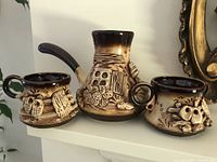 Photo showing the coffee/chocolate pot and two matching cups with embossed Ukrainian country fence and owl designs, in dark brown and beige glaze.