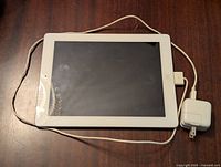Front view of white Apple iPad 2 16GB showing screen and connected charger cable with wall adapter on wooden surface.
