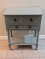 Front view of the antique grey console table showing three drawers, ring pull handles, turned legs, and lower shelf.
