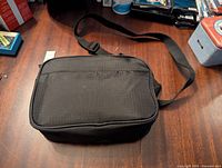 Front view of black nylon crossbody bag with top zipper and front pocket, flat on a wooden surface.