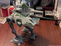 Side angled photo of the Star Wars AT-AP Walker toy showing detailed leg structure, turret, and cannon with various packaging and books in the background.