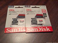 Photo showing front of two identical unopened SanDisk Ultra 256GB microSDXC cards with adapters in retail packaging, placed side by side on a wooden surface.