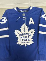 Front view of blue Toronto Maple Leafs hockey jersey with white maple leaf logo, number 34, and A alternate captain patch.