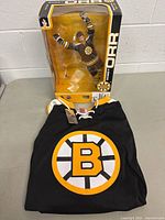 Photo showing black Boston Bruins hockey jersey with yellow and white Bruins logo, folded on table, with Bobby Orr action figure in original box behind it.