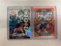 Photo shows two 2021 Justin Fields rookie football cards in protective plastic sleeves. One card is a colorful Panini Certified with 2021 large text and holographic finish. The other card is a Prizm Orange parallel with cracked ice pattern and holographic shine.