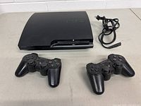 Top view of PS3 Slim console with two black DualShock 3 controllers and power cord to the upper right.