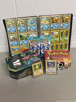 Open binder pages showing water and grass type Pokémon cards, with tin, card box, and sealed packs arranged in front