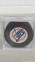 Front view of the hockey puck in protective case, showing Winnipeg Jets logo and Ed Olczyk signature in silver marker.