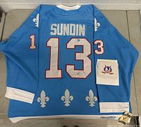 Light blue CCM hockey jersey laid flat showing full back with 'SUNDIN' nameplate and red-trimmed white number '13', fleur-de-lis logos along hem and sleeves, and A.J Authentic certificate envelope to side.
