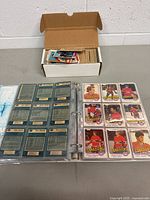 Open cardboard box containing loose vintage hockey cards and a hockey card binder shown with cards inside clear sleeves featuring player photos on front and stats on back.