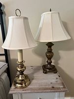 Photo showing two table lamps made of brass with different base designs and lampshades, positioned on a wooden surface against a wall.