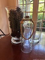 Photo showing three glass hurricanes: a tall flared one containing pinecones, a medium cylindrical one empty, and a bottle-shaped vase with a cork and rope handle
