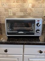 Front view of a Black & Decker toaster oven with two control dials, sitting on a kitchen counter with stone backsplash.