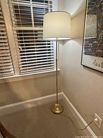 Photo of a tall brass standing lamp with adjustable height, topped with a white drum lampshade, placed on carpeted floor near window and framed artwork.