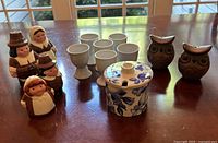 Photo shows two pairs of ceramic salt and pepper shakers, six white egg cups, and one floral jam pot with lid arranged on wooden surface under window light.