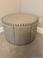 Full view of the round beige linen ottoman with brass nailhead trim placed against wall on carpeted floor.