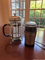 Photo showing a glass French press with metal frame and black handle next to a Capresso electric coffee grinder with stainless steel body and clear plastic lid, both on a wooden table by a window.