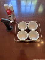 Photo showing crème brûlée set components including four white ramekins on a metal rack, black kitchen torch, and two fuel cartridges on wooden surface with window light.