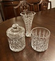 Photo showing a flared cut crystal vase and two cut crystal candy jars on wooden table in front of antique style wooden furniture.