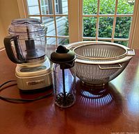 Photo shows Cuisinart mini food processor, Pampered Chef Measure Mix & Pour container with whisk inside, and three white colanders on a wooden table near window.