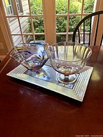 Side view of the three glass items on wooden table by window: multicolored ruffled hand blown bowl, clear footed bowl with ridges, and rectangular decorative tray.