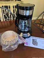 Black Hamilton Beach 12 cup coffee maker on a table with glass carafe in place, packaged coffee filters, a plastic scoop, and instruction manual visible.