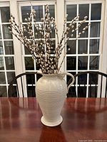 Photo of large cream colored ceramic decorative pitcher with handle and horizontal ridges on a wooden table with chairs in the background.