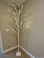 Photo of an 8-foot tall faux birch tree with small white lights on branches, placed in the corner of a beige-colored room on a white square base.