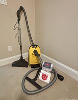 Photo of yellow Miele canister vacuum cleaner with attached hose and floor nozzle. Additional hose coiled in front with vacuum bags shown beside it.