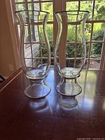 Two large clear glass hurricane candle holders standing on a table in front of a window with garden view.
