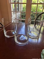 Three clear glass hurricane candle holders, about 8 inches tall, placed on a wooden table in front of a window with outdoor greenery visible.