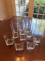 Photo of 7 tall square bottom highball glasses and 4 shorter square bottom whiskey glasses arranged on wooden table near window with garden view.
