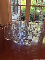 Photo showing thirteen stemless wine glasses on table by window. Two larger glasses for red wine and eleven smaller for white wine including etched glasses.