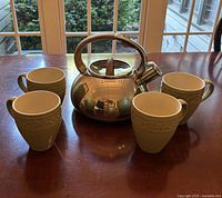 Photo showing stainless steel whistling kettle with a teak handle and knob in the center, surrounded by four matching light beige ceramic tea mugs with embossed decorative patterns.