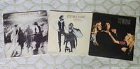 Three Fleetwood Mac vinyl records including Live, Rumours, and Mirage albums shown side by side on a patterned surface.