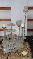 Various sizes of silver toned crystal beaded pillar and votive candle holders shown with 3-tiered round serving tray, rectangular mirrored serving tray, and small footed glasses on top.