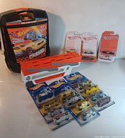 Photo showing the Hot Wheels branded case, semi truck car trailer, and 18 Hot Wheels cars in original packaging along with loose loose vintage cars on a white background