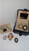 Black leather jewelry box with mirror inside lid, several cameo brooches and pins displayed inside and outside the box