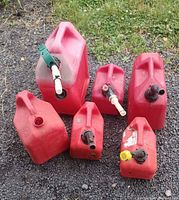 Photo showing six various size red plastic gas cans grouped on gravel and grass. Five have spouts, one is missing the spout.