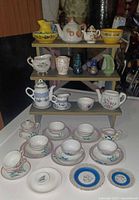Three-tier wooden stand holding multiple miniature tea sets including tea pots, cups, saucers, small bowls, and decorative vases arranged in groups.