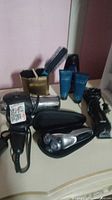Photo shows men's personal care items including electric razor, Wahl hair clippers, blow dryer, brush, two skin care tubes, and toothbrush holder on surface.