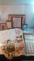 Wide view of quilting books, cross-stitch pattern booklets, framed cross-stitch art, and quilt pieces laid on table.