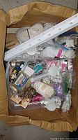 Cardboard box full of assorted sewing supplies including spools of thread, white elastics, plastic trims, needles, and small accessories