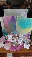 Photo showing two canvas paintings with abstract color designs, multiple white storage containers, decorative teal and purple glitter butterflies, and rocking horse figurines on a wooden table.